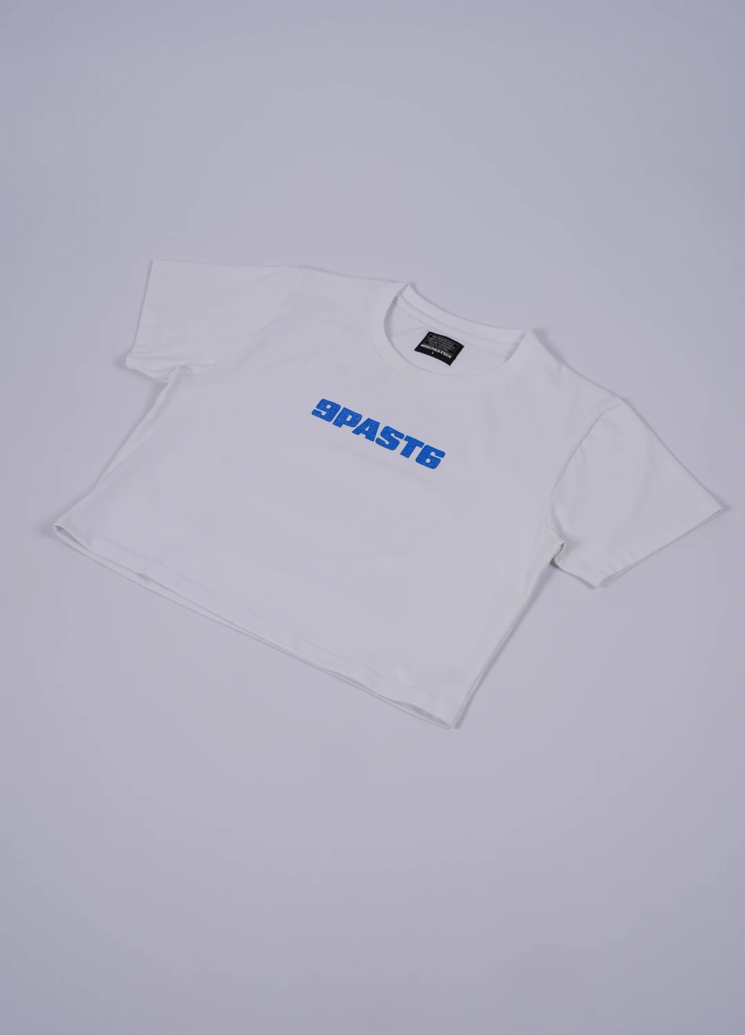 Real flat-lay photograph of a white cropped NinePastSix T-shirt with a blue 9PAST6 chest wordmark.