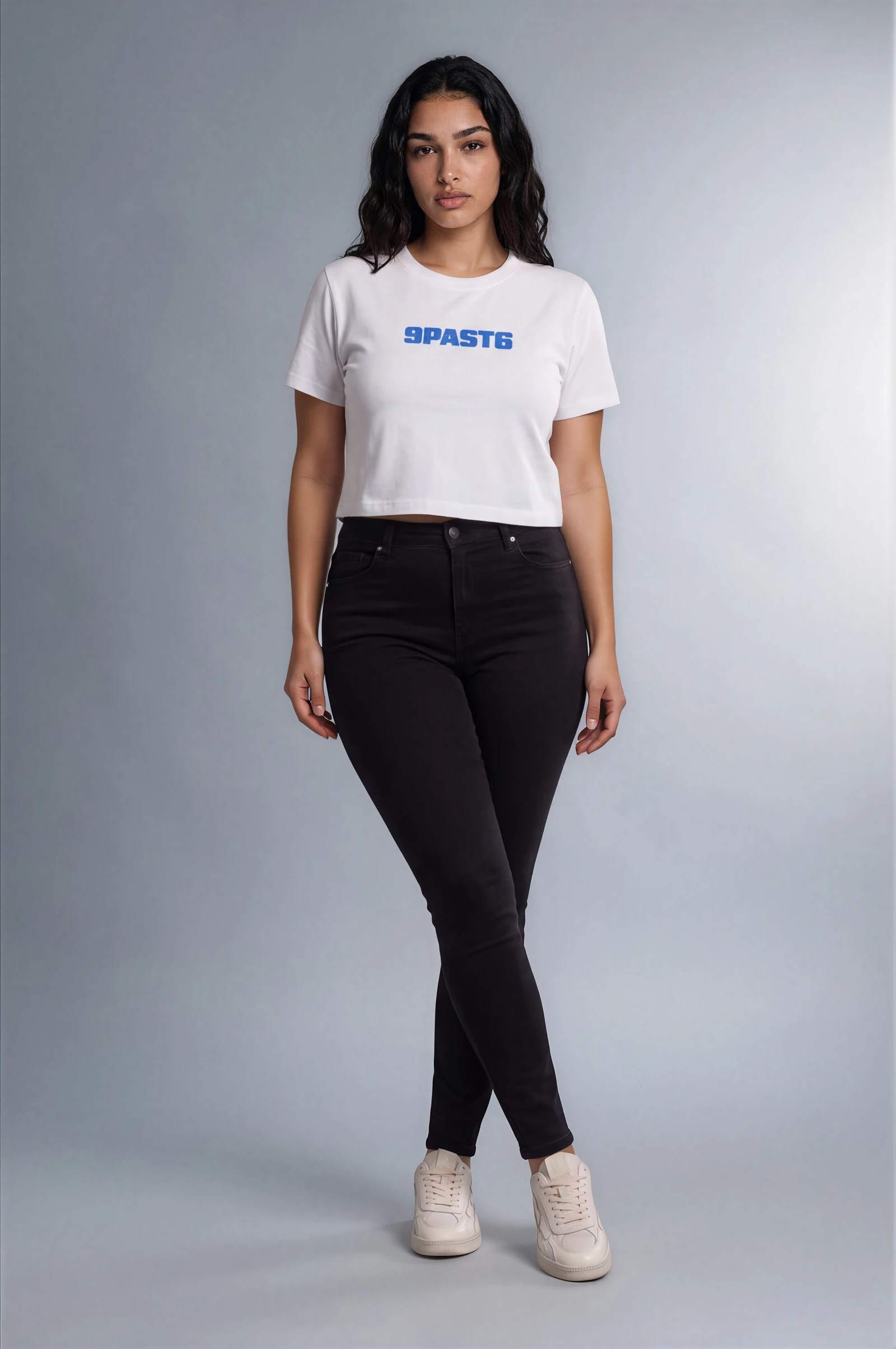 Full-length female model imagery styling the white blue-wordmark NinePastSix cropped T-shirt with black jeans and white sneakers.
